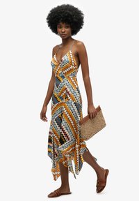 Vestido maxi de patrón de varios colores en chevrón, con un escote bajo, tirantes finos y dobladillo asimétrico. Lleva sandalias planas marrones y un bolso de mimbre.