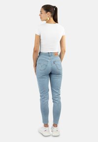 Helles blaues hoch-tailliertes Jeans mit einer passgenauen Form, einem Leder-Patch auf der Rückseite und weißen Sneakers. Das Model trägt ein schlichtes weißes Kurzarmhemd.