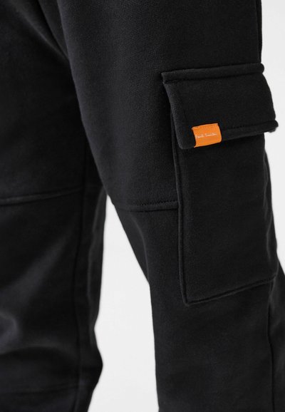 Mustat puuvillajoggersit, joissa on sivutasku ja oranssi etiketti. Sileä pinta, jossa on näkyviä ompelutietoja kankaassa.
