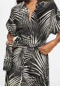 Blousejurk met knopen in zwart en crème met een palmblaadjespatroon. Heeft opgerolde korte mouwen en een stoffen ceintuur die in de taille is gestrikt.