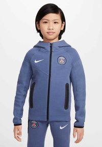 Veste à capuche zippée bleue avec capuche, poches zippées noires et logo du Paris Saint-Germain. Le matériau est doux avec une finition texturée.