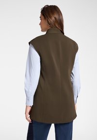 Olivově zelený bezrukávový blejzr s vysokým límcem, přiléhavý střih a ostrý silueta ramen, nošený přes světle modrou košili.