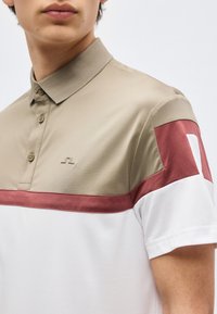 Νεαρό άτομο που φοράει κοντομάνικο πόλο μπλουζάκι με μπεζ επάνω μέρος, μπορντό ρίγα και λευκό κάτω μέρος, ορατό από τον λαιμό μέχρι το μέσο του κορμού.