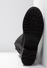 Botte en cuir noir avec une semelle épaisse et texturée tournée vers l'avant, reposant sur une surface blanche contre un mur blanc.