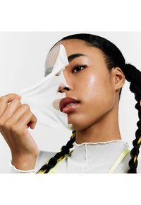 Femme aux cheveux tressés en train d’enlever un masque facial translucide, montrant une peau lisse en dessous, portant un haut blanc à col haut.