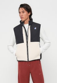 Gilet sans manches noir et crème en tissu synthétique avec une texture duveteuse. Il possède un col montant, une fermeture éclair à l'avant et un détail de logo.