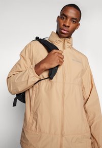 Veste légère beige avec fermeture éclair à l'avant, col montant, poignets élastiques et poches latérales. Présente un logo sur la poitrine et un tissu texturé.