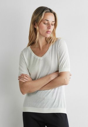 Blondynka ze skrzyżowanymi ramionami, ubrana w białą koszulkę z krótkim rękawem i dekoltem w serek oraz czarne spodnie na prostym, białym tle.