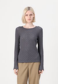 Blond bob-kihara juustega naine, kes kannab sobivat tekstuuriga tumedat halli pikkade varrukatega pluusi ja beeže laiade jalgadega pükse, seisab valge taustal.