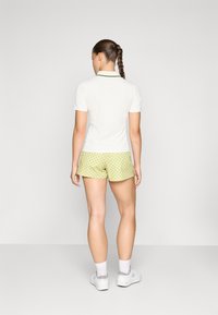 Polo majica v beli barvi z zelenimi detajli, ki je v paru z rumenimi kratkimi hlačami z pikastim vzorcem. Model stoji s konjskim repom in gleda stran.