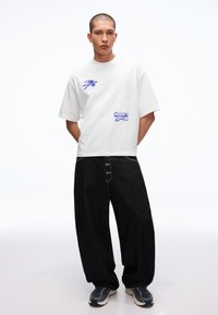 Fiatal férfi áll, hátra tett kézzel, fehér grafikás pólót, bő fekete gombos farmert és sötét tornacipőt visel fehér háttér előtt.