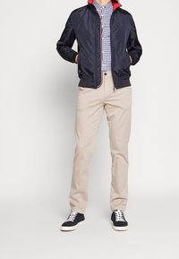 Marine bomberjack met rode kraag, geruite overhemd met knopen, beige chino's en marineblauwe sneakers met witte zolen. Glad, lichtgewicht materiaal.