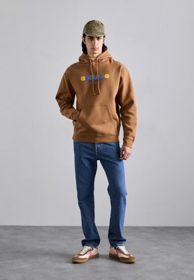 Hoodie marron avec une poche avant, comportant un texte bleu et jaune. Associé à un jean bleu clair et des baskets multicolores. La casquette est de couleur olive.