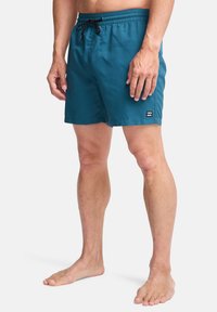 Bañadores de hombre en tejido verde azulado, con una cinturilla elástica y cordón negro, bolsillos laterales y un pequeño parche con el logo.