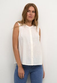 Blouse blanche sans manches avec un col rond, fermeture à boutons et texture subtile. Associée à un jean bleu ajusté.