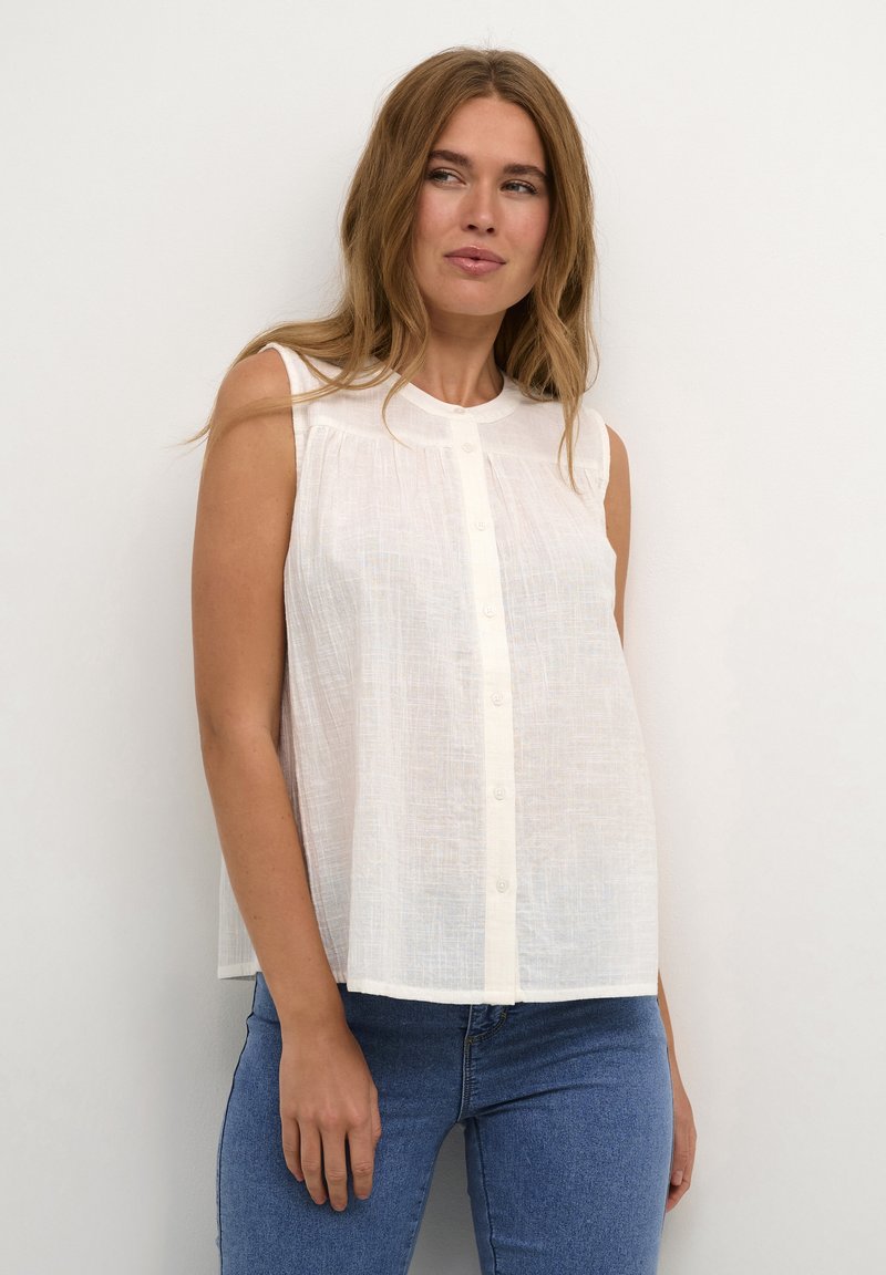 Hvid ærmeløs bluse med rund halsudskæring, knaplukning og subtil tekstur. Sammen med tætsiddende blå jeans.