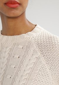 Nahaufnahme einer Person, die einen cremefarbenen, strukturierten Pullover mit Zopfmuster und roten Lippenstift trägt.