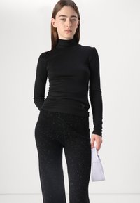 Črna dolgosleeve turtleneck majica v kombinaciji s črnimi hlačami z diskretnim bleščičastim vzorcem; model drži majhno sivkasto torbico.