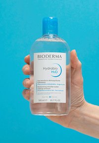 Una mano tiene una bottiglia da 500ml di Bioderma Hydrabio H2O, acqua micellare idratante per pelli sensibili, contro uno sfondo blu.