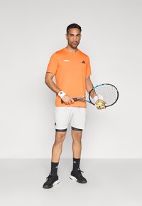 Πορτοκαλί κοντομάνικο μπλουζάκι με το λογότυπο "Tennis", λευκό σόρτς με σκούρες λεπτομέρειες, μαύρα παπούτσια του τένις, βραχιολάκια και ρακέτα που κρατά μια μπάλα του τένις.