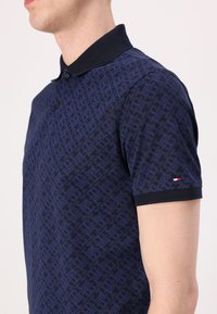 Marineblå polo-skjorte med geometrisk mønster, korte ermer, svart krage, to knapper, og en liten rød, hvit og blå logo på ermet.