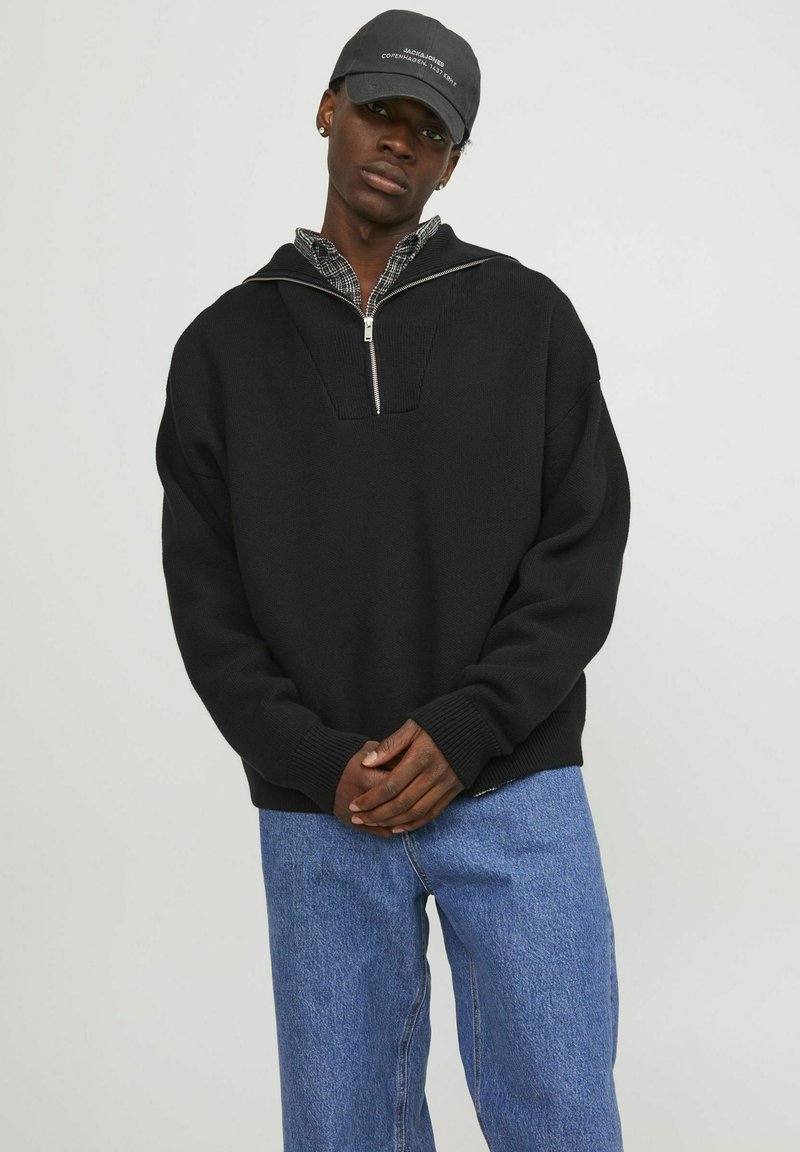 Schwarzer Pullover mit einem Viertel-Reißverschluss und gerippten Bündchen, über ein kariertes Hemd getragen. Hellblaue Jeans vervollständigen das Outfit.