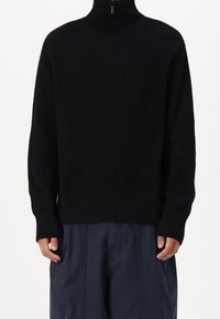 Jersey de punto acanalado negro con cuello alto y detalle de medio cierre, con mangas largas y un ajuste holgado. Llevado sobre pantalones azul oscuro.