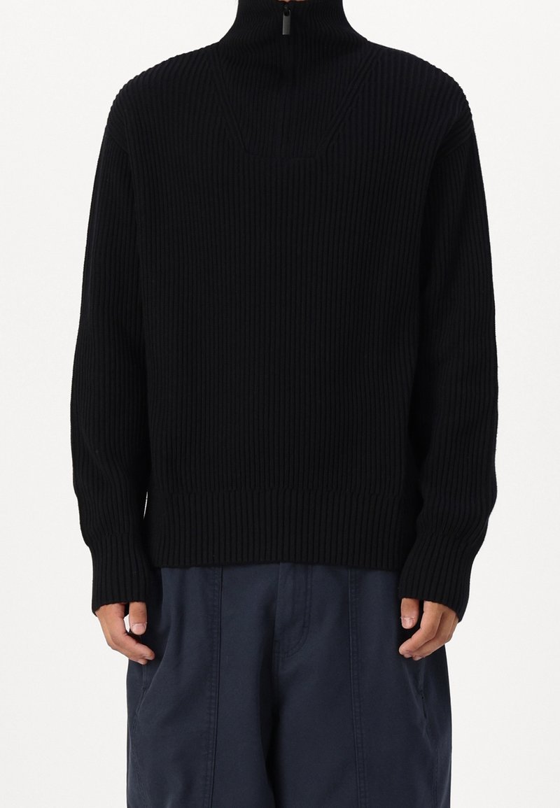 Jersey de punto acanalado negro con cuello alto y detalle de medio cierre, con mangas largas y un ajuste holgado. Llevado sobre pantalones azul oscuro.