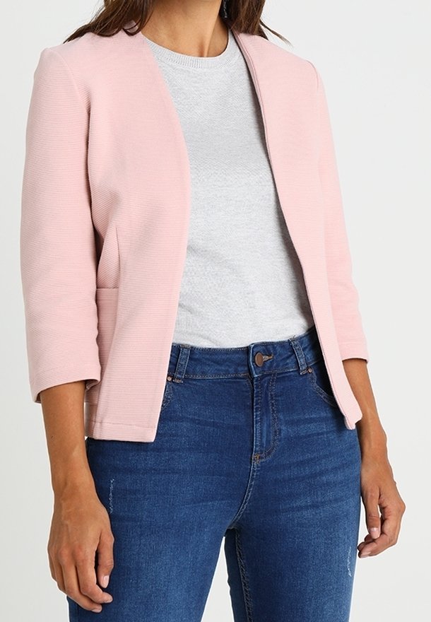 Femme portant un blazer court rose clair par-dessus un t-shirt gris à col rond et un jean bleu taille haute, debout devant un fond uni.
