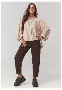 Beżowa, długa koszula z guzikami i bufiastymi rękawami, zestawiona z ciemnobrązowymi spodniami typu tapered oraz czarnymi butami na platformie. Gładka tekstura na całej powierzchni.