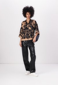 Sort blomstret bluse med beige detaljer, parret med sorte bukser i kunstlæder og hvide sneakers. Model poser foran en hvid baggrund.