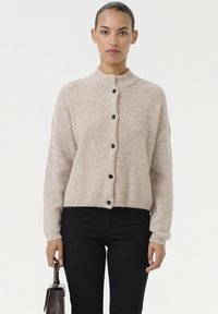 Beige geribde cardigan met een hoge kraag, zwarte knopen aan de voorkant en iets gekorte mouwen, gedragen met donkere spijkerbroeken.
