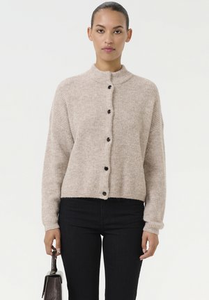 Beige geribde cardigan met een hoge kraag, zwarte knopen aan de voorkant en iets gekorte mouwen, gedragen met donkere spijkerbroeken.