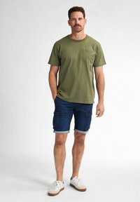 Olive green cotton t-shirt with crew neck and front pocket, paired with dark denim cargo shorts featuring rolled cuffs and side pockets. White sneakers.