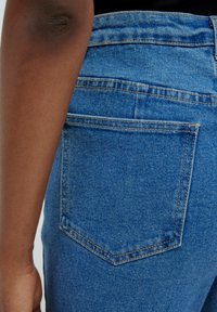 Primer plano de un brazo de piel oscura junto a un bolsillo trasero de mezclilla azul en unos pantalones vaqueros.