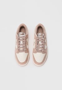 Ružové a béžové tenisky Nike s textúrovaným semišovým povrchom, perforovanou špičkou a plochými šnúrkami. Na boku sa nachádza biely Swoosh logo.