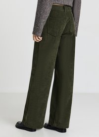 Pantalon large en velours côtelé vert foncé, présentant une texture nervurée verticale, deux poches arrière et une taille ajustée. Chaussures noires visibles.