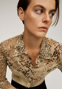 Mujer con cabello corto castaño, vistiendo una camisa de botones marrón con un patrón y un delicado collar de oro, mirando hacia la derecha contra un fondo simple.