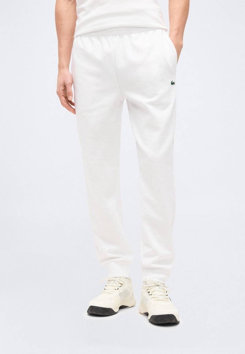 Λευκά παντελόνια jogger με ελαστική μέση και πλαϊνές τσέπες, φορεμένα με ανοιχτό μπεζ αθλητικά παπούτσια σε όρθιο άτομο.