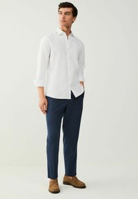 Chemise blanche à boutons, manches longues retroussées, associée à un pantalon slim-fit bleu marine et des mocassins marron. Design simple, texture de tissu nette.