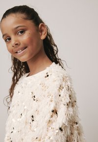 Fluffy white sweater with gold and black sequin accents, featuring a soft texture and round neckline, worn by a smiling girl with wavy hair.