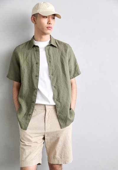 Chemise en lin olive à manches courtes sur un t-shirt blanc, assortie à un short beige. Une casquette beige clair complète l'ensemble.