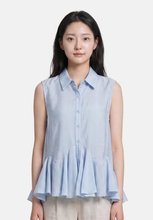 Giovane donna che indossa un top azzurro chiaro senza maniche con bottoni e orlo svasato a balze, in piedi davanti a uno sfondo bianco uniforme.