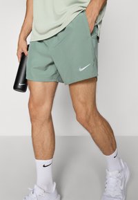 Shorts Nike verts avec une coupe décontractée, dotés d'une poche latérale et d'un logo discret. Portés avec des chaussures et des chaussettes blanches.