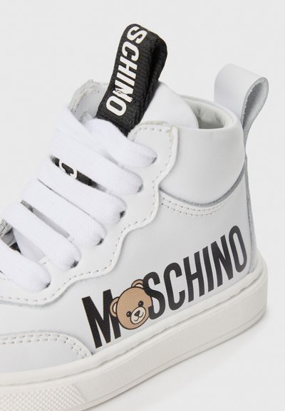 Witte leren hoge sneakers met een zwart logo etiket, witte veters en een bruine berenafbeelding op de zijkant. Textuur rubberen zool.