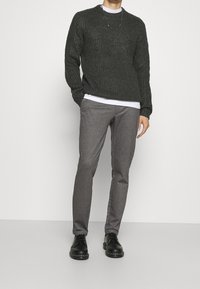Pull en tricot vert foncé avec une texture côtelée, superposé sur une chemise blanche. Un pantalon à motifs gris et des chaussures noires à lacets complètent le look.