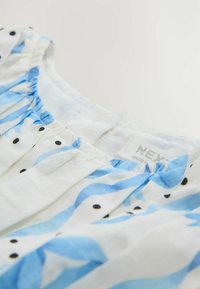 Biela blúzka s modrými vlnkovými vzormi a čiernymi bodkami; má naberaný výstrih a štítok s označením "NEXT." Ľahká bavlnená látka.