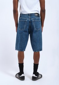 Pantalones cortos de mezclilla azul con un ajuste relajado, que cuentan con un diseño clásico de cinco bolsillos y un ligero desgaste, combinados con calcetines negros y zapatillas.