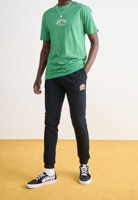 Camiseta de algodón verde con un logo circular, pantalones de chándal negros con un logo de color y zapatillas de cuadros en blanco y negro.