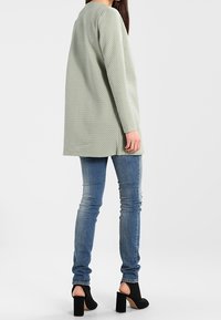 Světle zelená texturovaná bunda s dlouhým rukávem a uvolněným střihem, kombinovaná s modrými skinny džíny a černými kotníčkovými botami na podpatku. Hladká látka.